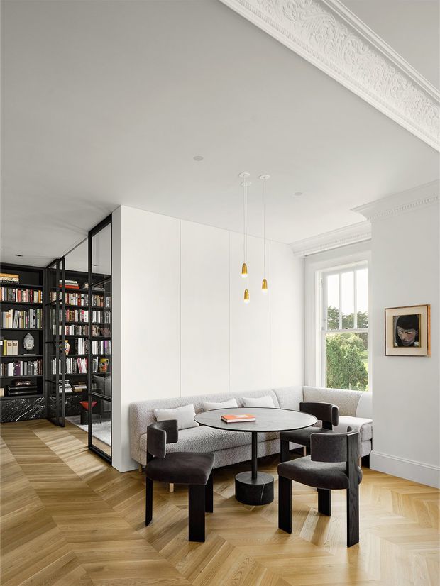 A refined sitting room with a pale upholstered curved sofa, round black coffee table, and several dark lounge chairs arranged on a light herringbone timber floor. A black-framed glazed bookcase or study zone is visible to the left, while tall windows and ornate white ceiling cornices add natural light and classical detail. Slim gold pendant lights and restrained artwork provide subtle accents.