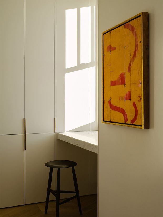 A warm, minimalist interior corner featuring tall pale built-in cabinetry with slim brass-toned handles, a light stone ledge or benchtop beneath a bright window opening, and a simple dark timber stool. A bold mustard-yellow and red abstract artwork in a dark wood frame hangs on the adjacent wall. Strong sunlight creates geometric shadows across the cabinetry and ledge, while dark timber flooring grounds the space.
