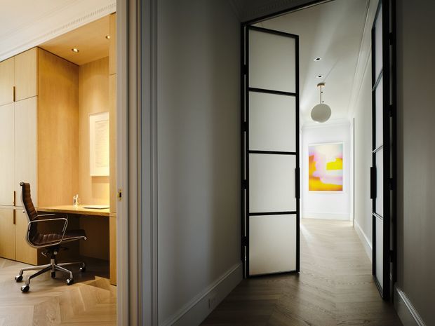 Interior view of a refined residential hallway with a black-framed glazed pivot door opening to a compact home office. The hallway has pale timber flooring, white walls with decorative cornices and skirting, recessed ceiling lights, and a spherical pendant. The office features warm timber built-in cabinetry, a desk, and a modern task chair. A colourful abstract artwork is visible at the end of the corridor.