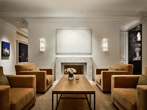 Symmetrical formal living room with four caramel-brown upholstered armchairs arranged around a rectangular timber coffee table. A marble-clad fireplace with a framed artwork above forms the central focal point, flanked by decorative wall sconces. Ornate crown moulding, ceiling detailing, dark timber flooring, and a black glass-backed bar or display cabinet contribute to a refined, classic interior.