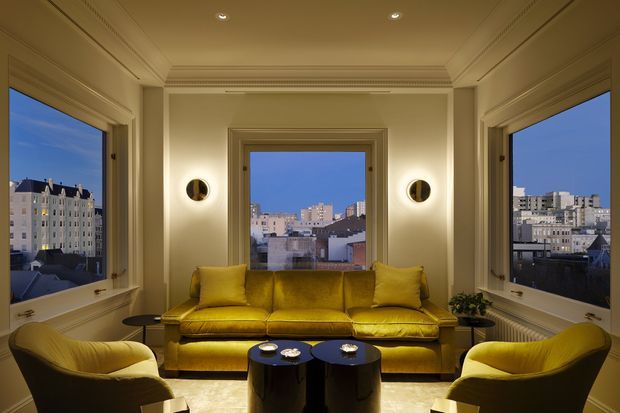 A refined living room with a symmetrical three-sided window bay overlooking a dense urban setting at dusk. The room features a mustard-yellow sofa and matching curved armchairs, dark round nesting coffee tables, illuminated circular wall sconces, cream painted wall panelling and moulded ceiling detailing. The composition is warm, polished and balanced, with a strong connection to the city view.