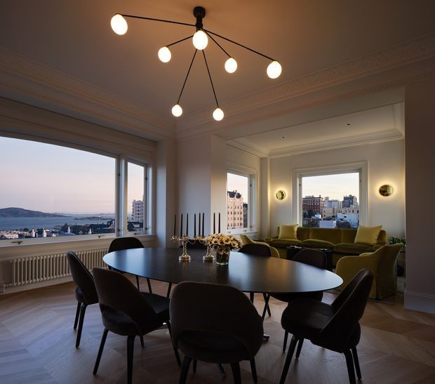 A refined dining and living area in a classic apartment, featuring a large dark oval dining table with upholstered dining chairs, an olive-green sofa and armchairs, decorative wall sconces, and a sculptural multi-arm pendant light. Tall windows provide expansive city and waterfront views. The room has ornate ceiling cornices, deep window reveals, white-painted walls, herringbone timber flooring, and a warm, elegant atmosphere.