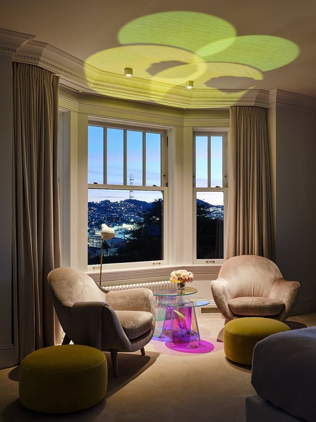 Elegant sitting room with a curved bay window overlooking a lit hillside cityscape. The room features traditional architectural detailing including ornate cornices and panelled trim, full-height taupe curtains, plush sculptural armchairs and ottomans, and a vivid transparent glass coffee table. A colourful projected light pattern creates a playful focal point across the ceiling and floor.