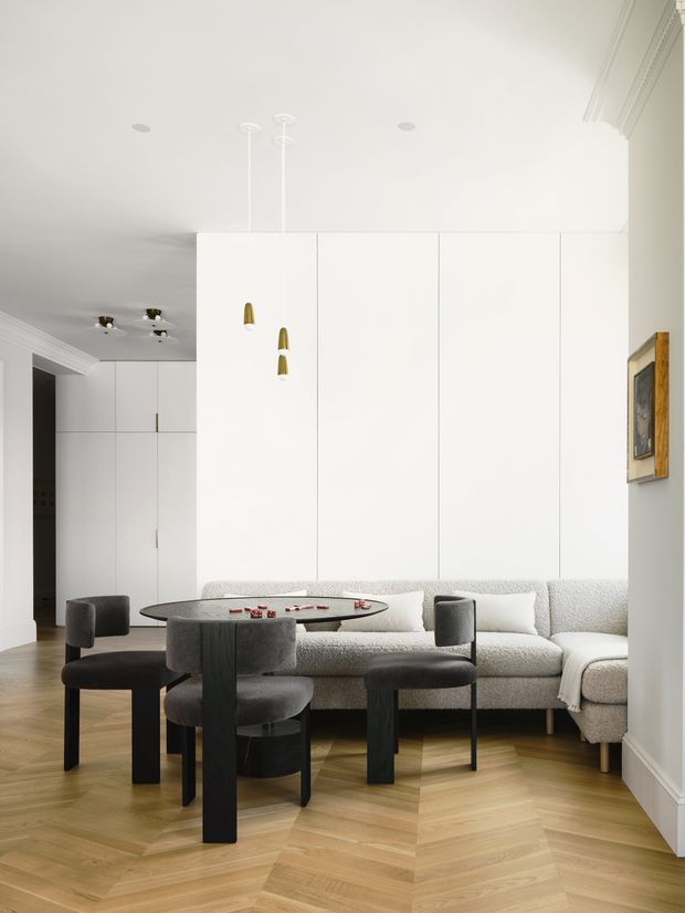 Bright, refined open-plan living and dining area with a large round dining table and dark upholstered chairs positioned in front of a textured light sofa. The room features white floor-to-ceiling built-in cabinetry or wall panelling, warm herringbone timber flooring, recessed ceiling lights, sculptural pendant lights, detailed cornices, and a framed artwork. The palette is predominantly white, cream, light timber, and charcoal grey, creating a clean contemporary-classic atmosphere.