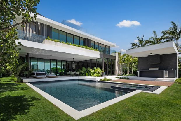 A contemporary two-storey residence with extensive floor-to-ceiling glazing, white flat roof slabs, dark vertical screens, and glass balustrades. The landscaped backyard features a rectangular swimming pool, timber pool deck, and a covered outdoor kitchen or entertaining pavilion with dark cabinetry and a wall-mounted screen. Lush tropical planting surrounds the clean-lined architecture.