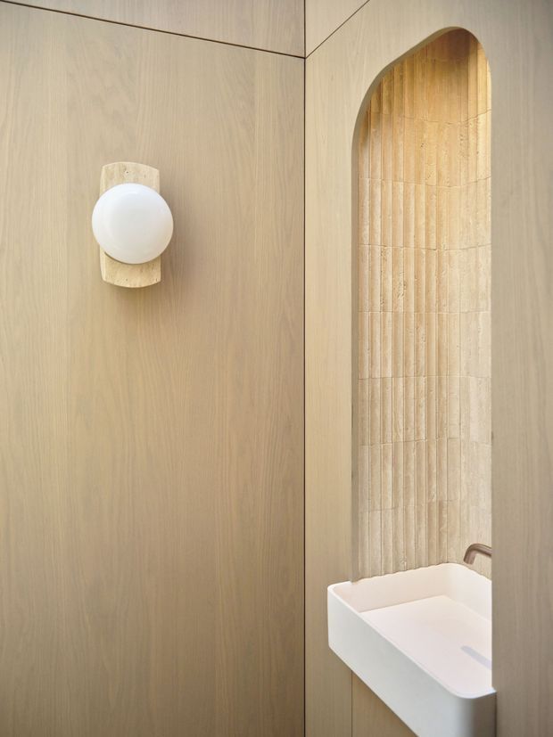 A refined compact bathroom interior featuring warm light timber wall panelling, a softly arched recessed niche lined with vertically fluted beige tile, a white wall-mounted basin, and a sculptural rounded wall sconce. The palette is warm and monochromatic with minimalist detailing.