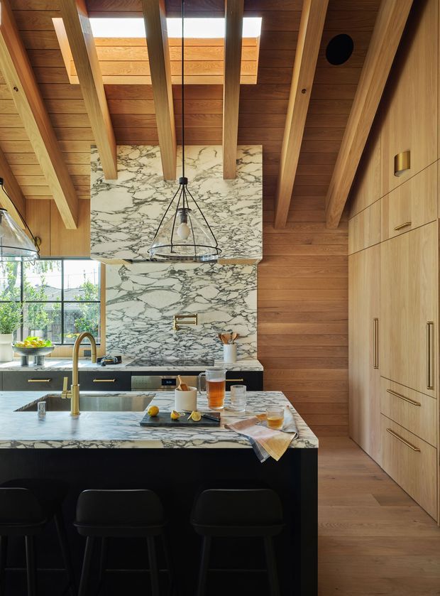 A sophisticated kitchen with a large marble island and matching marble splashback, dark lower cabinetry, and extensive warm timber cabinetry and wall finishes. The ceiling features exposed timber beams, timber lining, and multiple skylights that bring in natural light. A sculptural pendant hangs above the island, which has seating for three. Floor-to-ceiling integrated cabinetry lines the right wall, while a glazed exterior opening provides views to greenery outside.