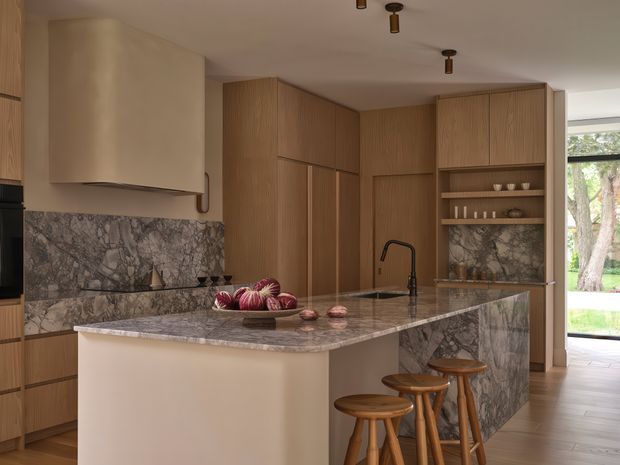 Warm, contemporary kitchen with extensive light timber cabinetry, a large marble-patterned stone island and matching splashback, integrated shelving, a sculptural rangehood, and three wooden stools at the island. The kitchen opens toward a glazed exterior doorway with greenery visible beyond.