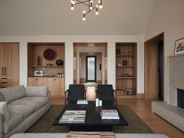 Warm, refined contemporary living room with a large textured sectional sofa, two dark lounge chairs, and a substantial black rectangular coffee table arranged on a muted area rug. Built-in timber cabinetry and open display shelving line the rear wall, framing a central hallway and dark-framed glazed entry door. A sculptural branching pendant light hangs from the high ceiling, while a pale stone or plaster fireplace surround anchors the right side of the room.