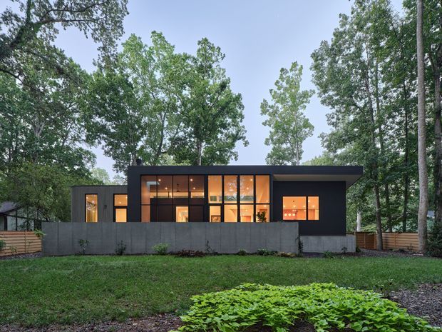 A contemporary single-storey residential house set among mature trees, viewed from the front across a landscaped lawn. The dark, rectilinear facade has a broad flat roof, extensive floor-to-ceiling glazing, warm amber interior lighting, and a dark solid privacy wall with timber fencing at the sides.