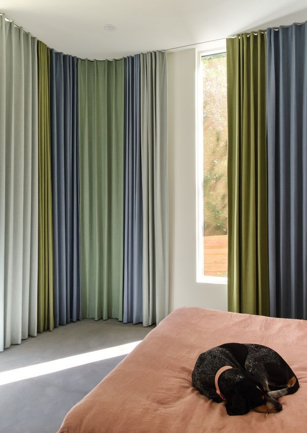 A bright bedroom corner with floor-to-ceiling pleated curtains in alternating sage green, olive, slate blue, grey, and white panels surrounding a corner window. A bed with a muted blush-pink cover occupies the foreground, with a sleeping black-and-tan dog on top. The room has a simple white ceiling, pale walls, and a light grey carpet, creating a colourful yet calm interior.