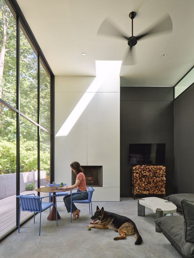 A bright, contemporary living room with floor-to-ceiling black-framed glazing along one wall, a sculptural ceiling fan, a central fireplace with integrated firewood storage, and a dark media wall. A small dining/work table and chairs sit beside the glazing, while a sectional sofa and sculptural coffee table occupy the foreground. Polished concrete flooring, strong natural light, and views of leafy vegetation create an open connection to the outdoors.