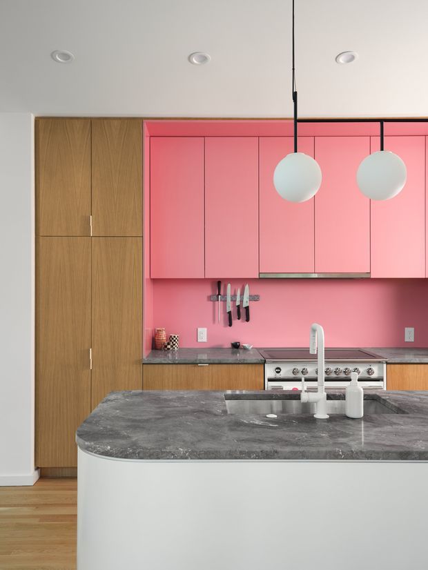 Contemporary kitchen with a vivid pink slab-front cabinetry wall, tall natural wood pantry cabinetry, dark grey stone countertops, and a large white island with an integrated sink and raised gooseneck faucet. Two spherical pendant lights hang above the island, while stainless steel appliances and a magnetic knife rack add functional detail. The space has a clean, playful, high-contrast finish with recessed ceiling lights and timber flooring.