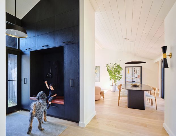 A split-view interior showing a dark, highly integrated entry and storage wall on the left, including a built-in bench nook, tall cabinetry, glazing, and pendant lighting, opening toward a bright open-plan dining area on the right. The dining space features a light timber floor and white timber-lined ceiling, a dark sculptural dining table, timber dining chairs, a display cabinet, pendant light, and wall sconce. A person and dog are present but are incidental to the architecture.