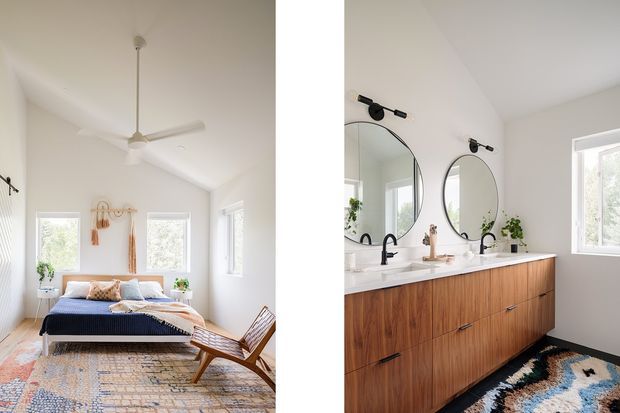 Bright split-view interior showing a serene bedroom on the left and an adjacent bathroom vanity on the right. The bedroom has white walls, a high sloped ceiling with a white ceiling fan, a low dark upholstered bed, woven rug, timber lounge chair, and restrained natural decor. The bathroom features a long timber vanity with white countertop, twin round mirrors, black wall-mounted lights and tapware, and a window providing natural light.