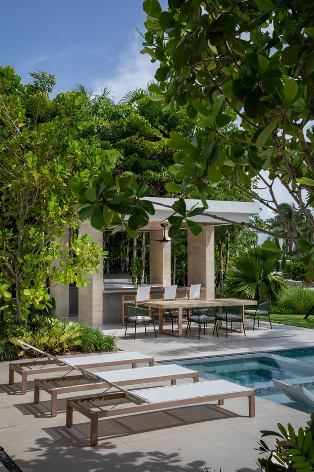A lush tropical outdoor pool area with a contemporary covered dining pavilion. The space features a rectangular swimming pool, pale paved decking, several timber-framed sun loungers, an outdoor dining table with chairs, integrated outdoor bench seating, stone-clad columns, a flat white roof, and dense tropical landscaping.