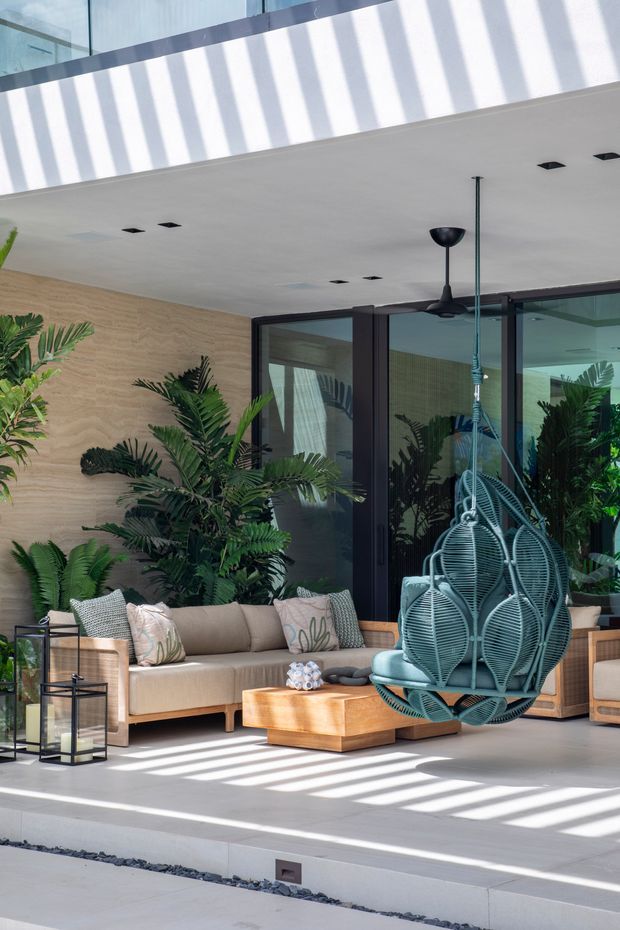 A bright, contemporary indoor-outdoor lounge space with a covered ceiling, full-height dark-framed glazing, textured warm timber-look wall cladding, and abundant tropical planting. The area is furnished with a neutral upholstered sofa, timber coffee table, lanterns, and a distinctive suspended woven egg chair. Strong linear sunlight and shadows fall across the pale floor, creating an airy resort-like atmosphere.