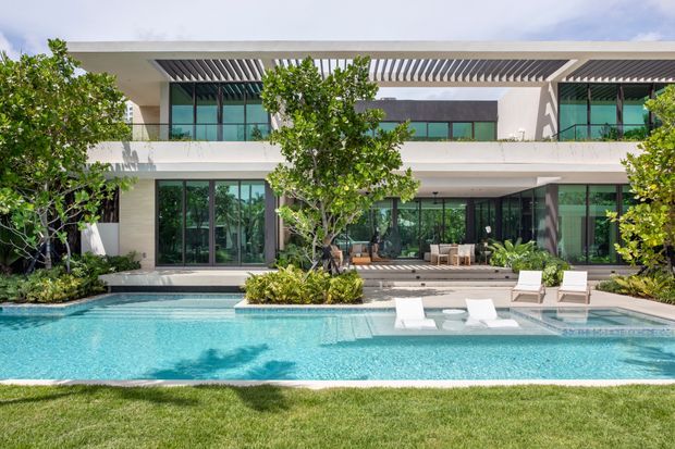 A bright, contemporary two-storey residence viewed from the landscaped backyard, with extensive floor-to-ceiling glazing, white rendered walls, deep horizontal overhangs and a slatted pergola. A large turquoise swimming pool with a shallow sun shelf, submerged loungers and surrounding planting dominates the foreground, while outdoor seating is visible beneath the covered terrace.