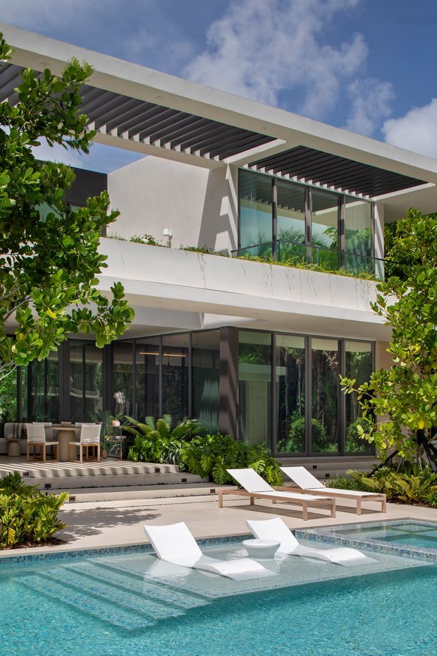 A bright contemporary multi-level house surrounds a landscaped swimming pool. The facade features white rendered walls, extensive dark-framed glazing, deep covered terraces, and prominent open pergolas with dark slatted sun-shading. Poolside loungers and tropical planting create a resort-like outdoor setting.