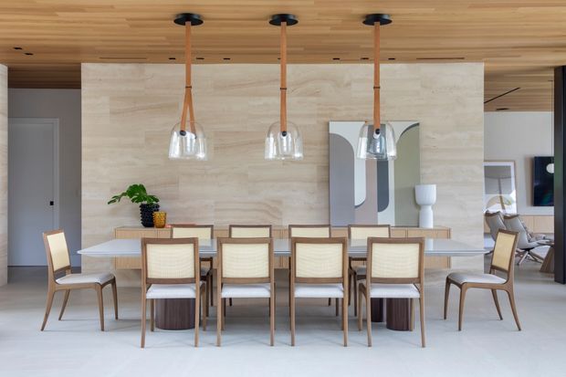 A refined contemporary dining area with a long pale dining table surrounded by upholstered timber dining chairs. The room features a full-height light stone feature wall, warm timber ceiling with recessed lighting, and three suspended glass pendant lights with timber-toned stems. A large abstract artwork, console decor, and glimpses of an adjacent living area complete the bright, neutral interior.