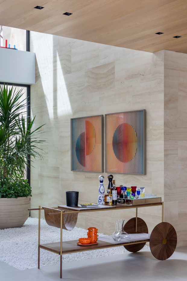 A bright, sophisticated interior vignette featuring a sculptural timber-and-metal bar console against large pale natural-stone walls. The console includes rounded timber details and shelves displaying glassware, bottles, and decorative objects. A large potted plant sits beside a black-framed window, while colourful geometric artwork hangs above the console. The ceiling is clad in light timber with recessed spotlights, and white decorative gravel surrounds the planting area.