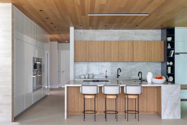 A spacious contemporary kitchen with a long island and three stools. The room features warm timber cabinetry and a timber-lined ceiling, pale stone wall and benchtop surfaces, integrated appliances, a dark open display shelf, and minimalist black tapware and lighting. A hallway and additional built-in storage extend beyond the kitchen.