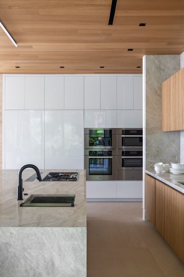 A refined contemporary kitchen with a large pale stone island, integrated dark sink and black gooseneck tap, flush gas cooktop, and extensive glossy white handleless cabinetry. A bank of stainless-steel ovens is set into the rear cabinetry. Warm timber lining spans the ceiling, while vertically fluted timber cabinetry and a matching stone benchtop feature along the right side. Large-format neutral flooring and a textured stone wall complete the clean, high-end fit-out.