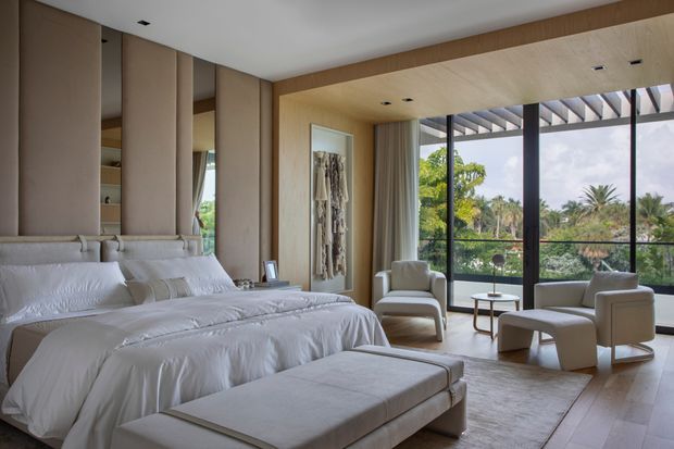 A refined contemporary bedroom with a large upholstered bed, layered neutral bedding, a matching upholstered bench, and a pair of sculptural lounge chairs beside a small round table. Full-height black-framed glazing opens the room visually to lush greenery, while warm timber wall and ceiling lining, soft beige upholstered panels, recessed lighting, and a textured wall hanging create a calm, luxurious retreat.