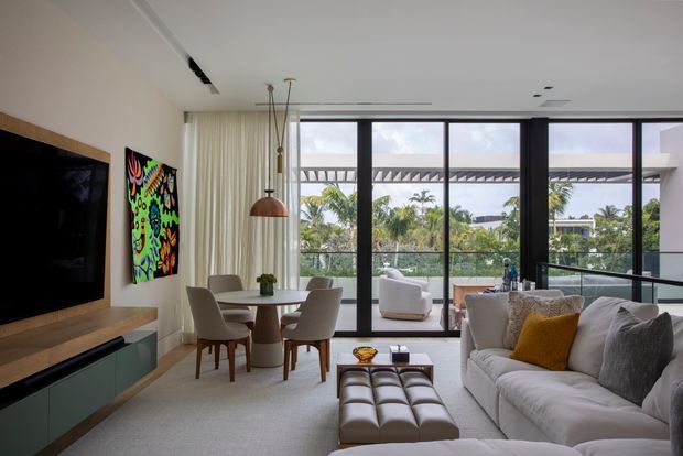Bright contemporary open-plan living and dining area with a large white sectional sofa, tufted ottoman, round dining table, upholstered dining chairs, wall-mounted television, low built-in media cabinetry, and colourful artwork. Floor-to-ceiling black-framed glazing opens to a balcony or terrace and overlooks lush tropical landscaping. A linear recessed ceiling light and suspended pendant add modern detailing.