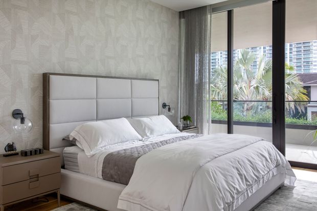 A bright, contemporary bedroom with a large upholstered bed and padded geometric headboard, layered white bedding, and a grey throw. The room features light patterned wallpaper, streamlined bedside joinery, globe wall sconces, sheer curtains, and full-height dark-framed glazing opening toward a planted outdoor terrace and city buildings.