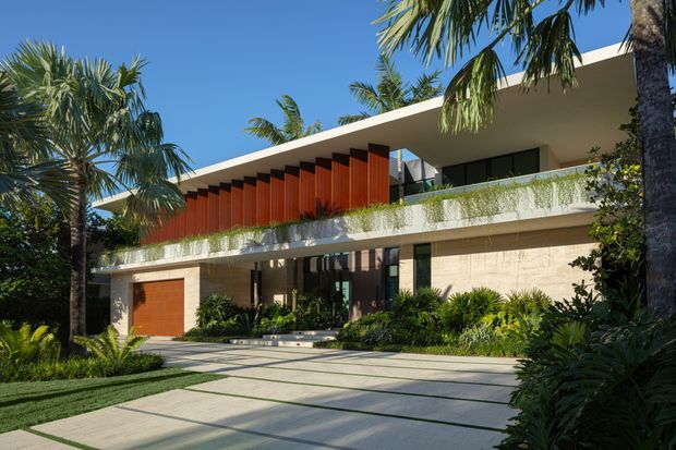 A contemporary two-storey tropical residence viewed from the front, featuring a broad flat roof, deep overhangs, exposed warm timber-toned vertical screening, pale textured masonry walls, extensive glazing, and a planted balcony edge. A large paved driveway and lush palm-filled landscaping frame the facade.