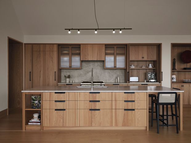 A warm, contemporary kitchen centred on a large timber island with integrated drawers and open shelving. The cabinetry extends across the rear wall, with glazed upper cupboards, a textured stone-look splashback, undermount sink, cooktop, and dark bar stools. Timber flooring, muted walls, and a linear track light create a refined, cohesive interior.