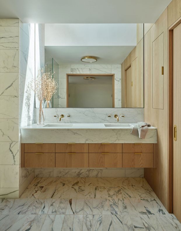 A refined contemporary bathroom featuring full-height veined marble wall and floor surfaces, a wide double vanity with a white marble benchtop and integrated basins, warm timber drawer cabinetry, brass tapware, a large frameless mirror, and a timber door with recessed panel detailing. Natural light enters from the left, creating a calm, luxurious atmosphere.