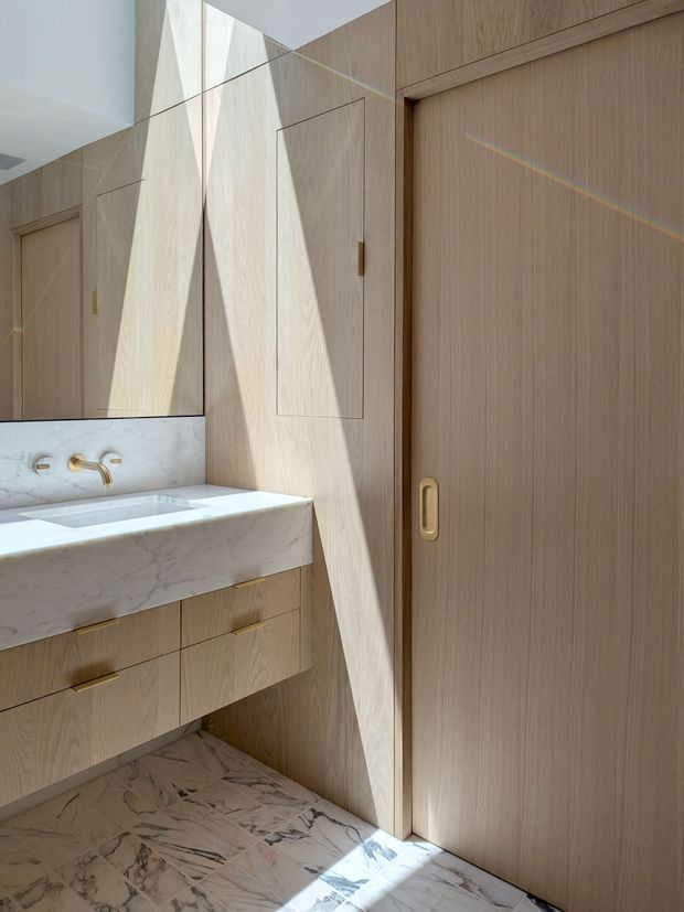 A refined bathroom with pale timber veneer cabinetry, a marble vanity and integrated basin, brass tapware and hardware, and a large mirrored wall. A timber sliding door and concealed storage panels continue the warm, minimalist joinery, while strong natural sunlight creates geometric highlights across the surfaces.