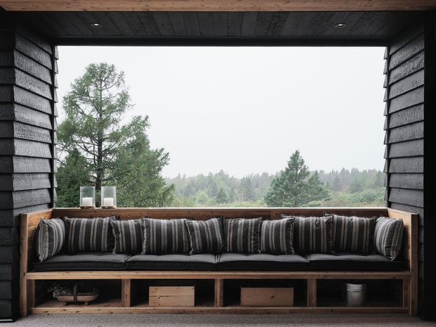 A covered outdoor lounge nook with a long built-in timber bench, dark striped cushions, and open views toward a wooded landscape. The space is framed by dark textured cladding and a timber ceiling, with integrated open storage below the seating.