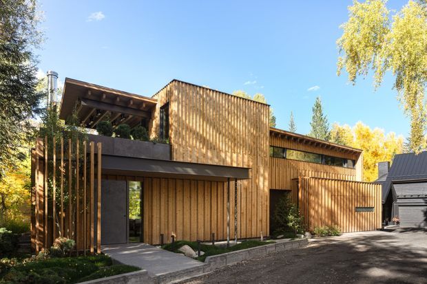 A contemporary multi-level residence viewed from the street, featuring extensive vertical timber cladding, dark metal accents, deep roof overhangs, recessed glazing, and a partially screened upper terrace. The home sits within a leafy, established landscape and includes a broad driveway and adjacent dark-clad structure.