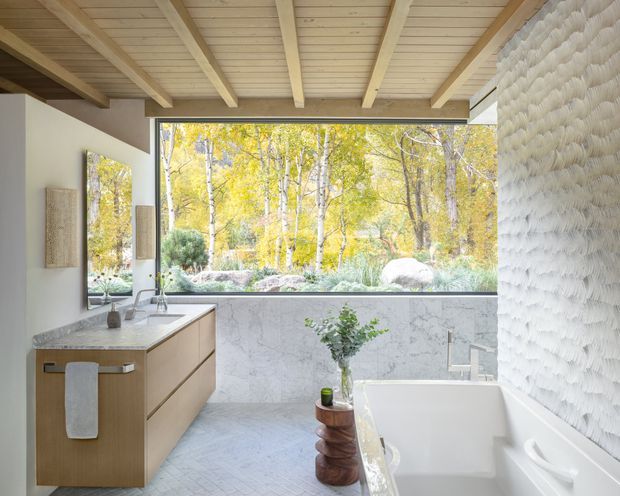 Bright, refined bathroom with a floating light-wood vanity, pale stone surfaces, a freestanding white bathtub, and a large picture window overlooking a wooded landscape. The ceiling has exposed light timber beams and boards, while the right wall features textured sculptural white tile or plaster. Natural light and organic materials create a serene, spa-like atmosphere.