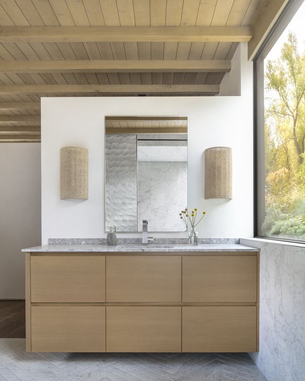 A refined bathroom vanity area with a wide floating light-wood drawer unit, pale veined stone benchtop, integrated basin and wall-mounted tapware. Above the vanity is a large textured mirror or mirrored feature panel, flanked by woven cylindrical wall sconces. The room has a striking exposed light timber beam-and-board ceiling, white plaster walls, marble-look wall cladding, and a large black-framed window overlooking trees. The overall design is warm, calm, and contemporary with natural materials.