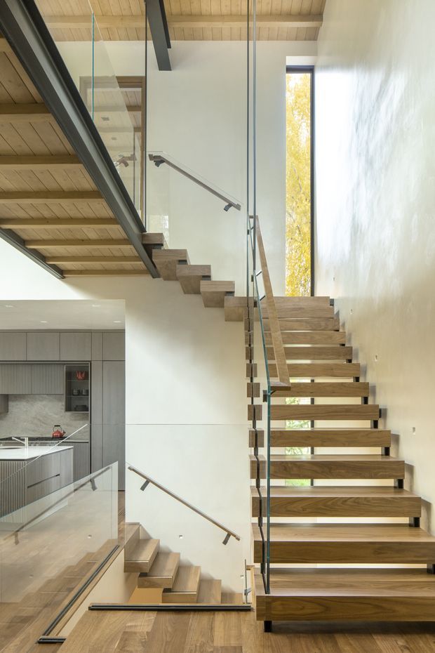 Interior view of a contemporary open-riser timber staircase with glass balustrades and slim metal handrails. The stair rises through a tall, light-filled void beside a modern kitchen, with warm timber ceiling boards, exposed dark structural beams, white plaster walls, and a narrow vertical window bringing in foliage-filtered daylight.