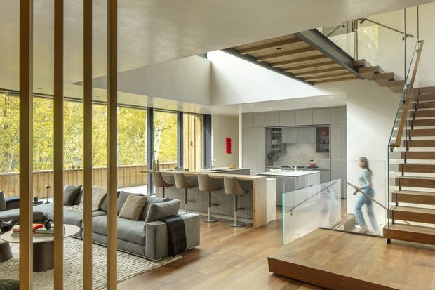 Open-plan contemporary home interior combining a living room, kitchen and dining area beneath a dramatic staircase and upper-level void. Floor-to-ceiling glazing brings in abundant natural light and views of trees, while warm timber flooring, vertical timber screens, a large island with bar seating, and a glass stair balustrade define the space.