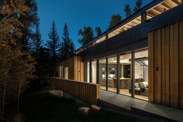 Twilight exterior view of a contemporary timber-clad house set among mature trees. The long, low building has extensive floor-to-ceiling glazing, a dark projecting upper roof or parapet, warm exposed timber soffits, and a raised deck or terrace. Interior kitchen and living areas are visible through the glazed doors, with a stair and furnishings illuminated inside.