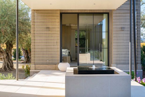 A bright contemporary outdoor courtyard or terrace framed by horizontal timber screening and large floor-to-ceiling glass doors. A rectangular dark water feature or reflecting pool sits in front of a pale stone or concrete plinth, with a partially visible refined interior beyond the glazing. Mature landscaping surrounds the space, and slim dark structural posts and a flat roof create a clean, minimalist architectural composition.