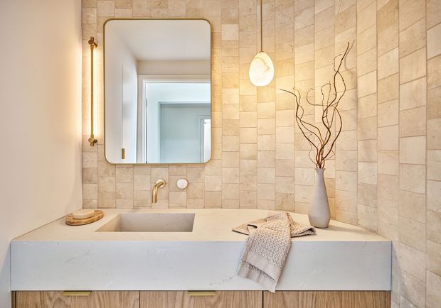 Contemporary bathroom vanity with a large pale stone or porcelain tiled feature wall, integrated rectangular basin, pale benchtop, timber-look cabinetry, brass tapware, rounded-edge wall mirror, and warm decorative lighting. Neutral beige and cream tones create a refined, softly lit atmosphere.