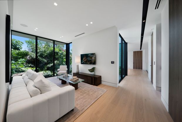 Bright contemporary open-plan living room viewed from a connecting hallway, featuring a large cream sectional sofa, timber flooring, a low dark timber media console, neutral rug, and expansive black-framed corner glazing overlooking greenery. A clean white ceiling with recessed lighting and linear air vents contributes to the minimalist architectural character. The hallway extends toward a timber door and additional rooms.