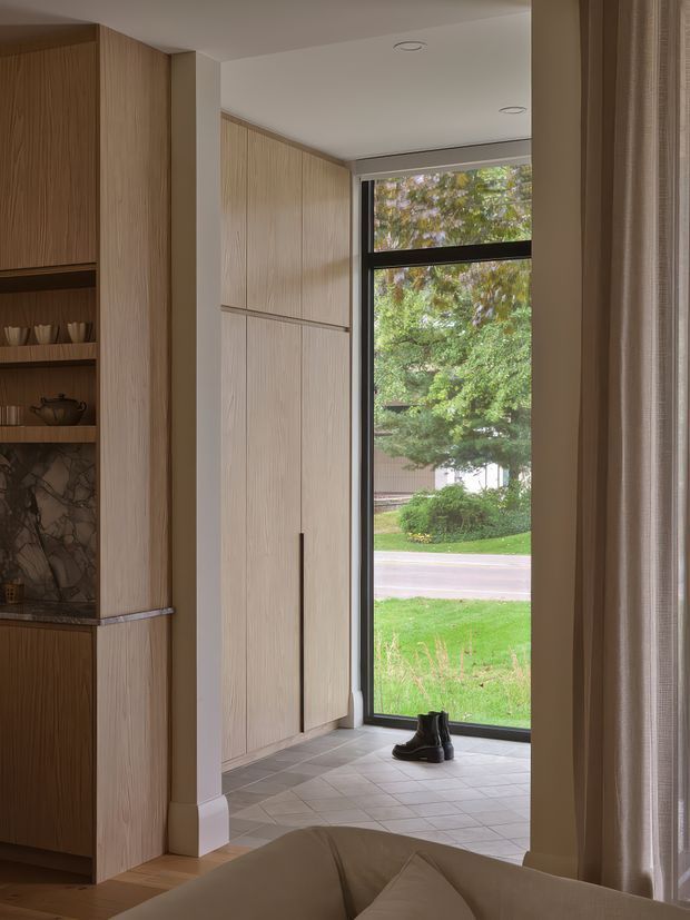 A refined residential entryway with floor-to-ceiling light timber built-in cabinetry, open display shelving, and a dark stone feature backing. Large black-framed glazing brings in views of greenery, while pale square floor tiles and neutral curtains create a calm, warm interior. A pair of boots near the window reinforces the entrance function.