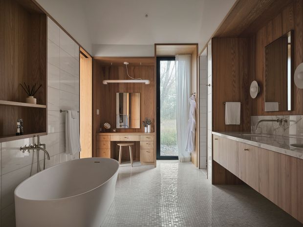 A spacious, refined bathroom with a freestanding oval bathtub in the foreground, a double vanity with marble-look benchtop and large mirror on the right, and a wall-mounted basin beneath open timber shelving on the left. Warm timber wall panelling and cabinetry contrast with pale tiled walls and a small-format light mosaic floor. A timber-framed glazed door brings in natural light, while a built-in timber dressing or storage area is visible at the rear.