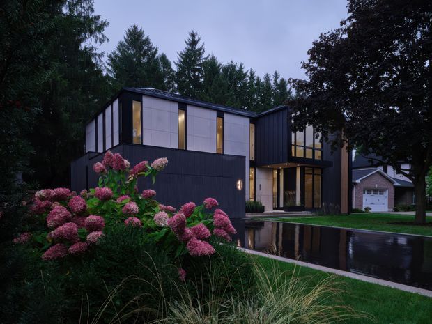 Exterior view of a contemporary multi-level residence at dusk, set among mature trees and landscaped gardens. The house features dark vertical cladding, pale rectangular wall panels, extensive warm-lit glazing, a projecting upper volume, and a flat or very low-pitched roof. A wet driveway reflects the building and surrounding lights, with pink hydrangeas and ornamental grasses in the foreground. A neighbouring garage is visible in the background.