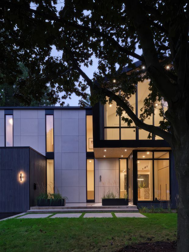 Contemporary multi-level home exterior viewed from a landscaped front yard at dusk. The facade combines pale grey panel cladding, dark vertical cladding and black-framed floor-to-ceiling glazing, with warm interior and recessed exterior lighting. A mature tree partially screens the building, while concrete stepping slabs, planted beds and lawn define the entry approach.