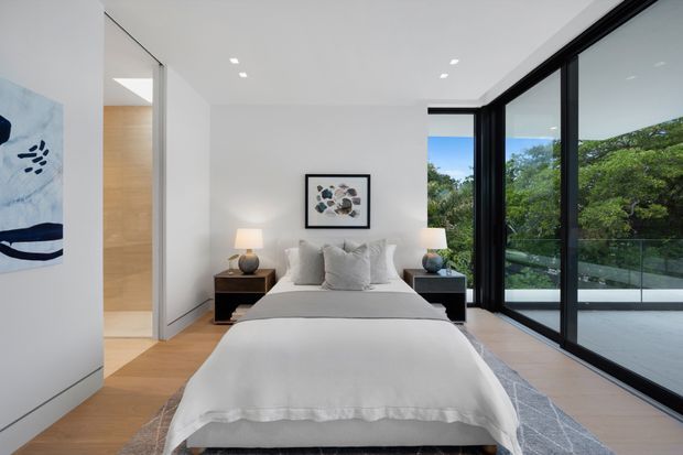Bright contemporary bedroom with a low-profile upholstered bed dressed in white linens, matching dark bedside tables and lamps, pale timber flooring, minimalist white walls, and large black-framed floor-to-ceiling glazing opening onto a balcony or terrace with greenery beyond. A framed abstract artwork and built-in wall joinery add subtle detail.