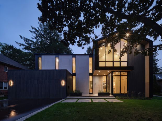 Twilight exterior view of a contemporary multi-level residence with a dark vertically ribbed facade, pale rectangular wall panels, extensive warm-lit glazing, and a projecting upper-floor volume. The house is set in a suburban landscaped lot with a paved driveway and front path.
